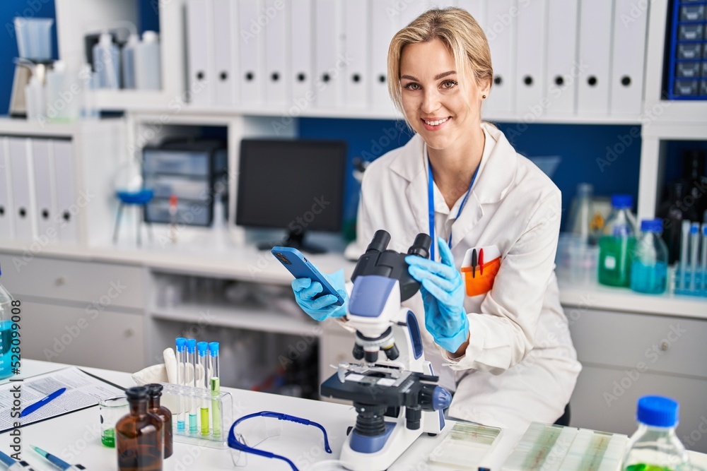 Young blonde woman scientist using microscope and smartphone at laboratory