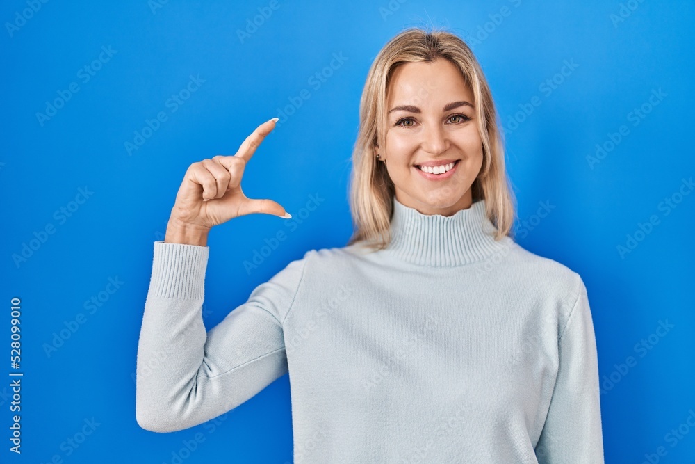 Young caucasian woman standing over blue background smiling and confident gesturing with hand doing small size sign with fingers looking and the camera. measure concept. Young caucasian woman standing over blue background smiling and confident gesturing with hand doing small size sign with fingers looking and the camera. measure concept.
