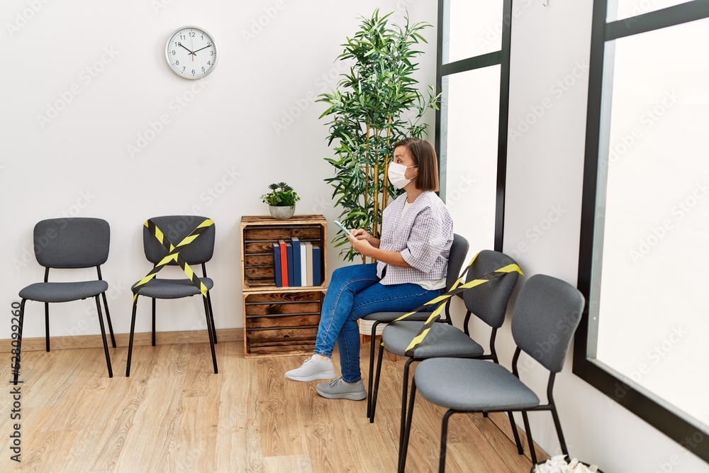 Middle age hispanic woman wearing safety mask using smartphone at waiting room Middle age hispanic woman wearing safety mask using smartphone at waiting room