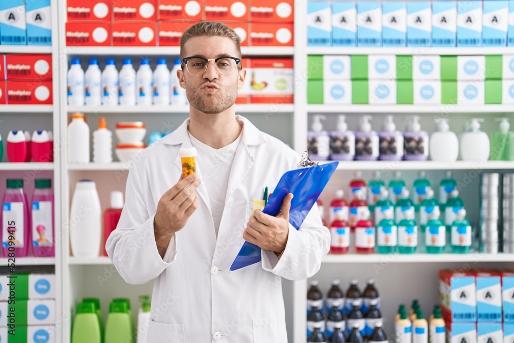 Young caucasian man working at pharmacy drugstore holding pills looking at the camera blowing a kiss being lovely and sexy. love expression. Young caucasian man working at pharmacy drugstore holding pills looking at the camera blowing a kiss being lovely and sexy. love expression.