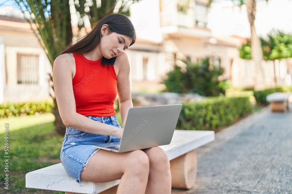 Young caucasian woman using laptop talking on smartphone sitting on bench at park Young caucasian woman using laptop talking on smartphone sitting on bench at park