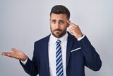 Handsome hispanic man wearing suit and tie confused and annoyed with open palm showing copy space and pointing finger to forehead. think about it.
