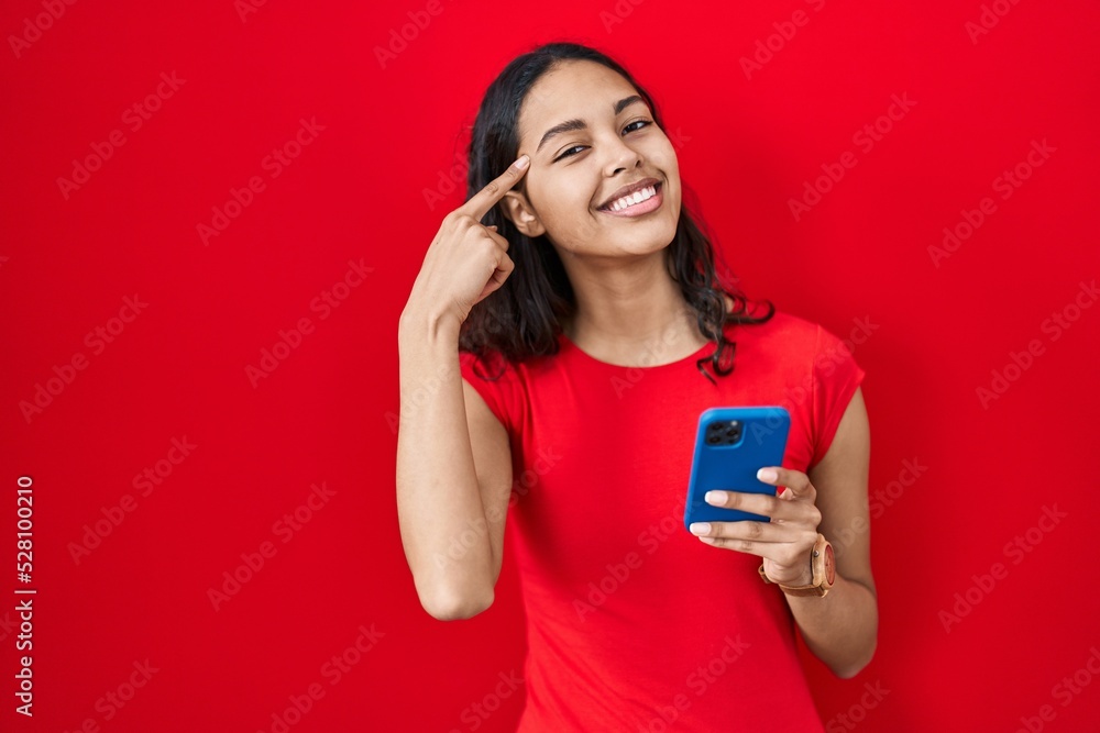 Young brazilian woman using smartphone over red background smiling pointing to head with one finger, great idea or thought, good memory Young brazilian woman using smartphone over red background smiling pointing to head with one finger, great idea or thought, good memory