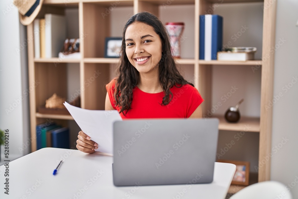 Young african american woman using laptop reading document sitting on table at home Young african american woman using laptop reading document sitting on table at home