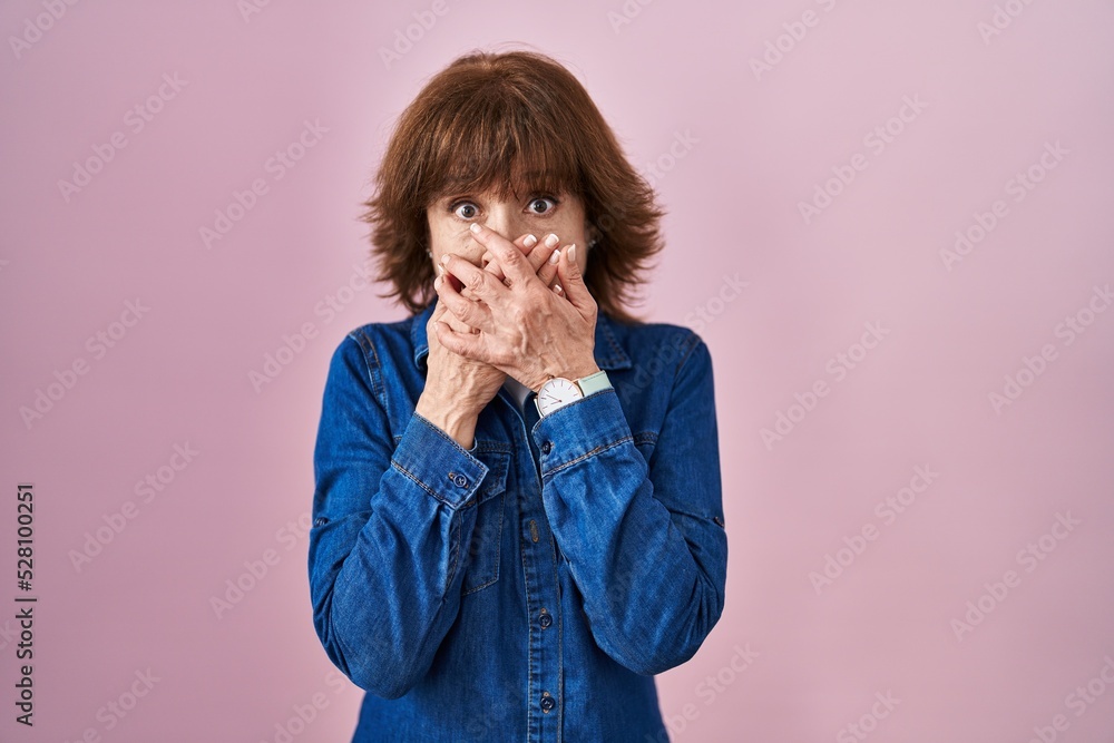 Middle age woman standing over pink background shocked covering mouth with hands for mistake. secret concept. Middle age woman standing over pink background shocked covering mouth with hands for mistake. secret concept.