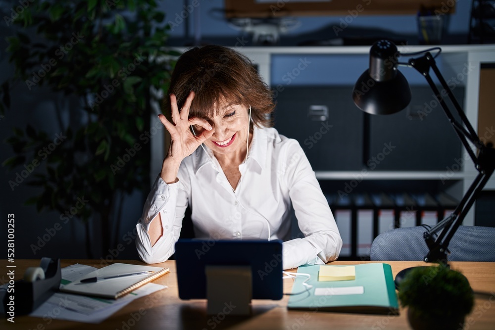 Middle age woman working at the office at night doing ok gesture with hand smiling, eye looking through fingers with happy face. Middle age woman working at the office at night doing ok gesture with hand smiling, eye looking through fingers with happy face.