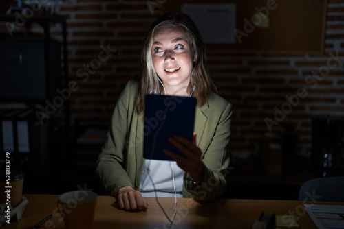 Blonde caucasian woman working at the office at night looking away to side with smile on face, natural expression. laughing confident.