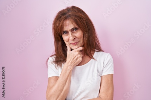 Middle age woman standing over pink background looking confident at the camera smiling with crossed arms and hand raised on chin. thinking positive.