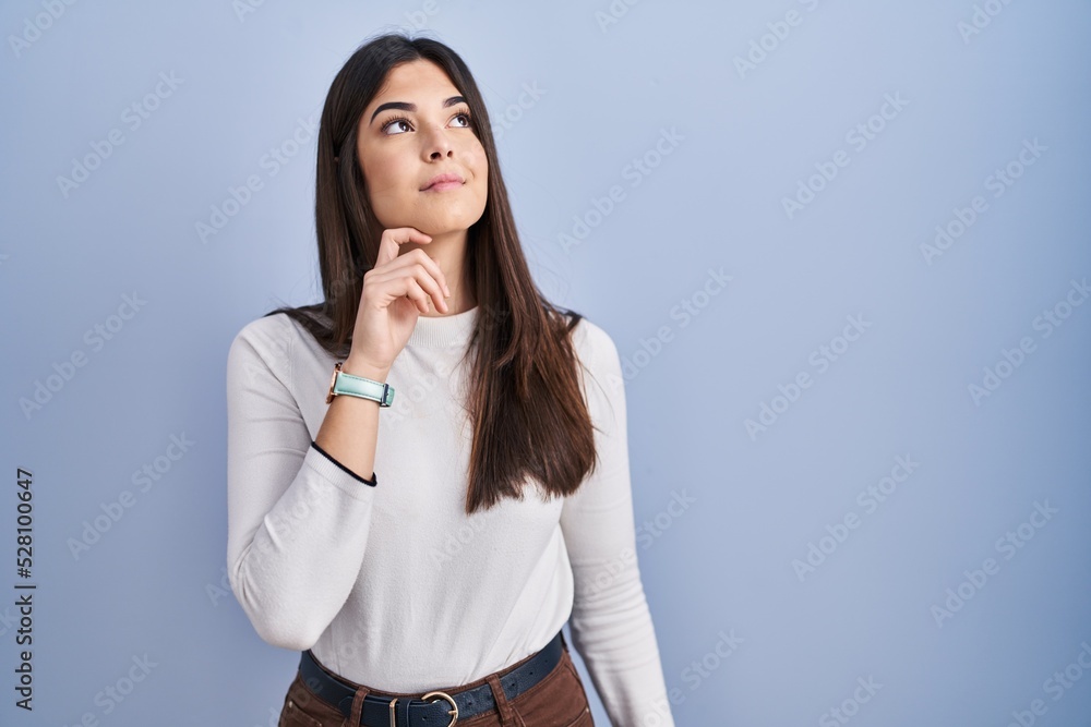 Young brunette woman standing over blue background with hand on chin thinking about question, pensive expression. smiling with thoughtful face. doubt concept. Young brunette woman standing over blue background with hand on chin thinking about question, pensive expression. smiling with thoughtful face. doubt concept.