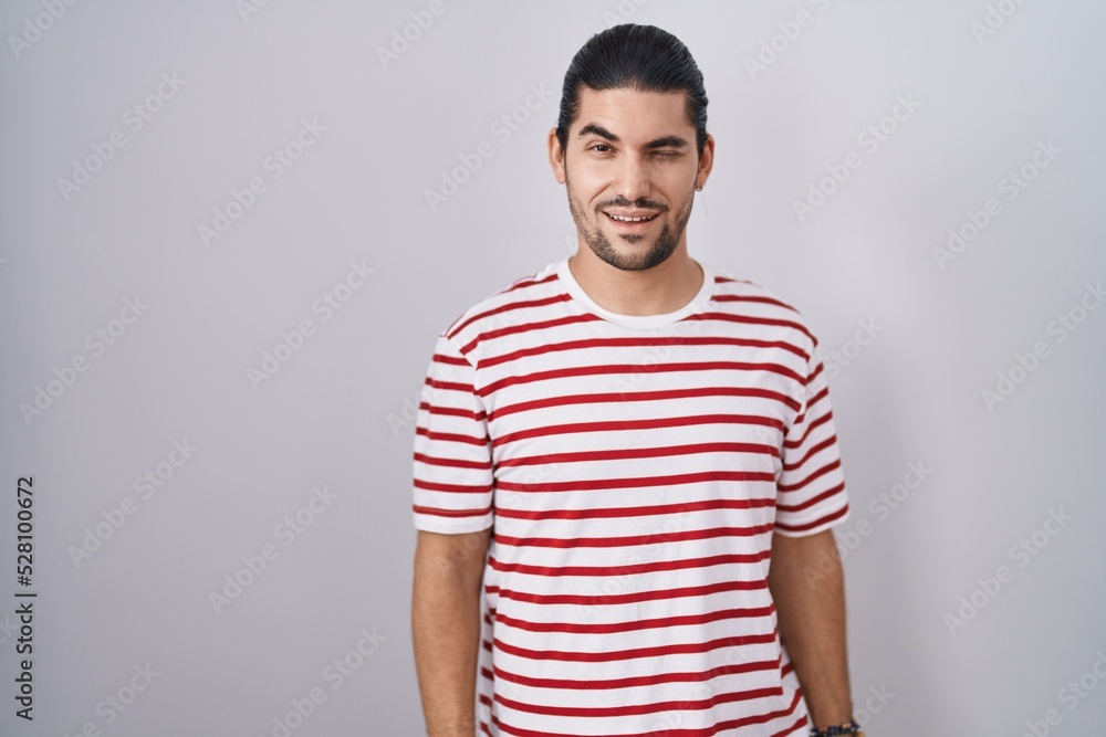 Hispanic man with long hair standing over isolated background winking looking at the camera with sexy expression, cheerful and happy face. Hispanic man with long hair standing over isolated background winking looking at the camera with sexy expression, cheerful and happy face.