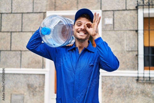 Young hispanic man holding a gallon bottle of water for delivery smiling happy doing ok sign with hand on eye looking through fingers photo