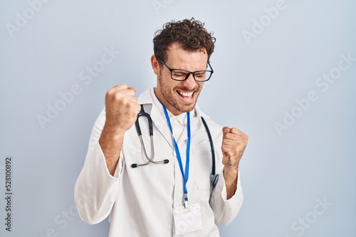 Young hispanic man wearing doctor uniform and stethoscope very happy and excited doing winner gesture with arms raised, smiling and screaming for success. celebration concept.