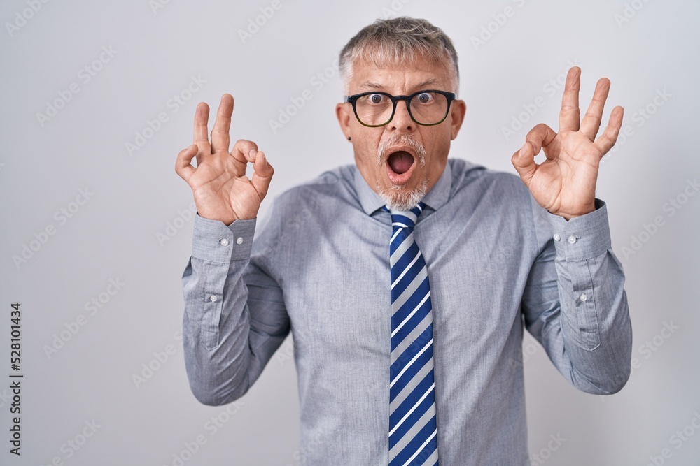 Hispanic business man with grey hair wearing glasses looking surprised and shocked doing ok approval symbol with fingers. crazy expression