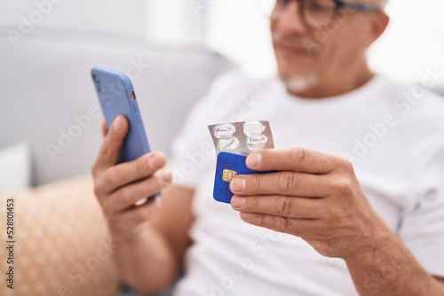 Middle age grey-haired man using smartphone and credit card holding pills at home