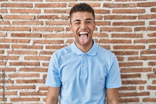 Brazilian young man standing over brick wall sticking tongue out happy with funny expression. emotion concept.