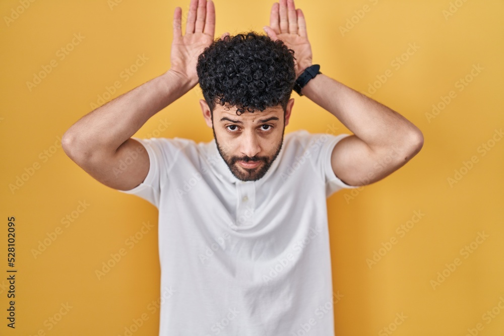 Arab man standing over yellow background doing bunny ears gesture with hands palms looking cynical and skeptical. easter rabbit concept. Arab man standing over yellow background doing bunny ears gesture with hands palms looking cynical and skeptical. easter rabbit concept.
