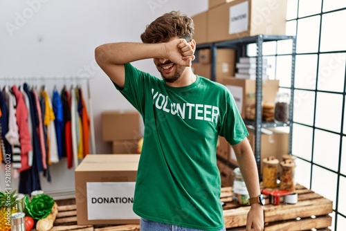 Young arab man wearing volunteer t shirt at donations stand smiling cheerful playing peek a boo with hands showing face. surprised and exited