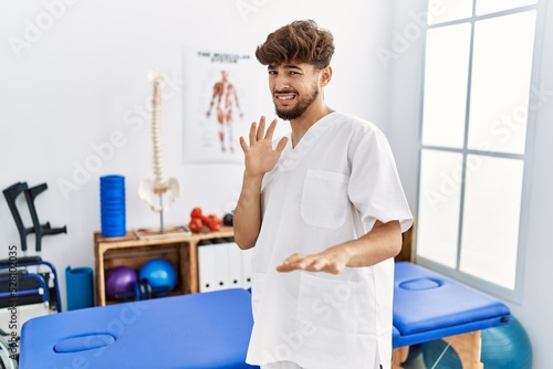 Young arab man working at pain recovery clinic disgusted expression  displeased and fearful doing disgust face because aversion reaction. with hands raised