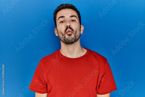 Young hispanic man with beard wearing red t shirt over blue background making fish face with lips, crazy and comical gesture. funny expression.