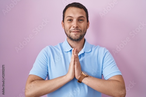 Hispanic man standing over pink background praying with hands together asking for forgiveness smiling confident. © Krakenimages.com