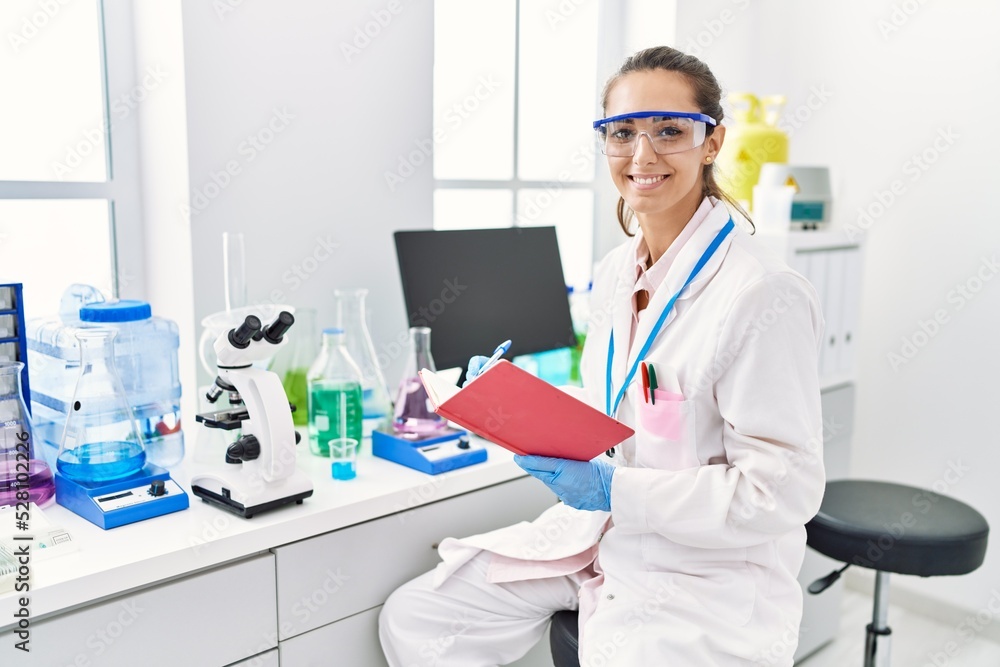 Young hispanic woman wearing scientist uniform writing on notebook using microscope at laboratory Young hispanic woman wearing scientist uniform writing on notebook using microscope at laboratory
