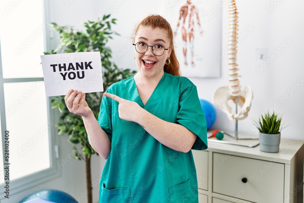 Young redhead physiotherapist woman working at pain recovery clinic holding thank you banner smiling happy pointing with hand and finger Young redhead physiotherapist woman working at pain recovery clinic holding thank you banner smiling happy pointing with hand and finger