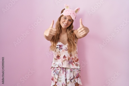 Young caucasian woman wearing sleep mask and pajama approving doing positive gesture with hand, thumbs up smiling and happy for success. winner gesture.