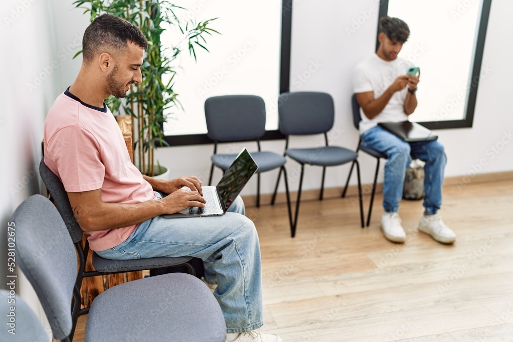 Two hispanic men using laptop and smartphone sitting on chair at waiting room