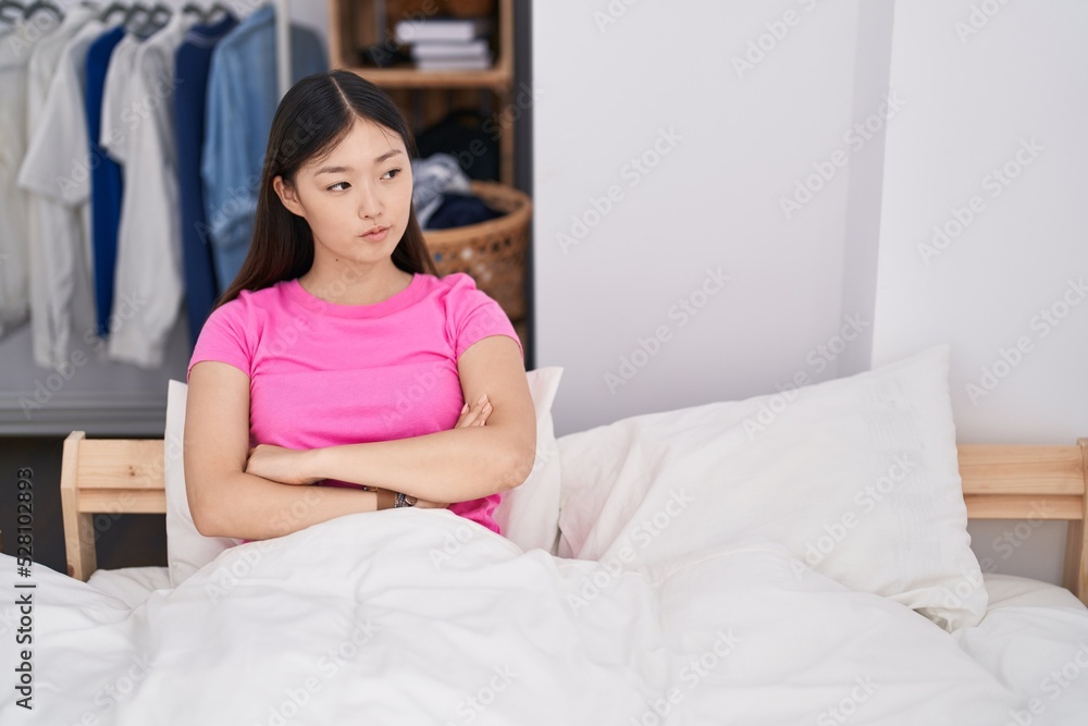 Chinese woman sitting on bed with arms crossed gesture and serious expression at bedroom Chinese woman sitting on bed with arms crossed gesture and serious expression at bedroom