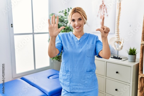 Beautiful blonde physiotherapist woman working at pain recovery clinic showing and pointing up with fingers number seven while smiling confident and happy.