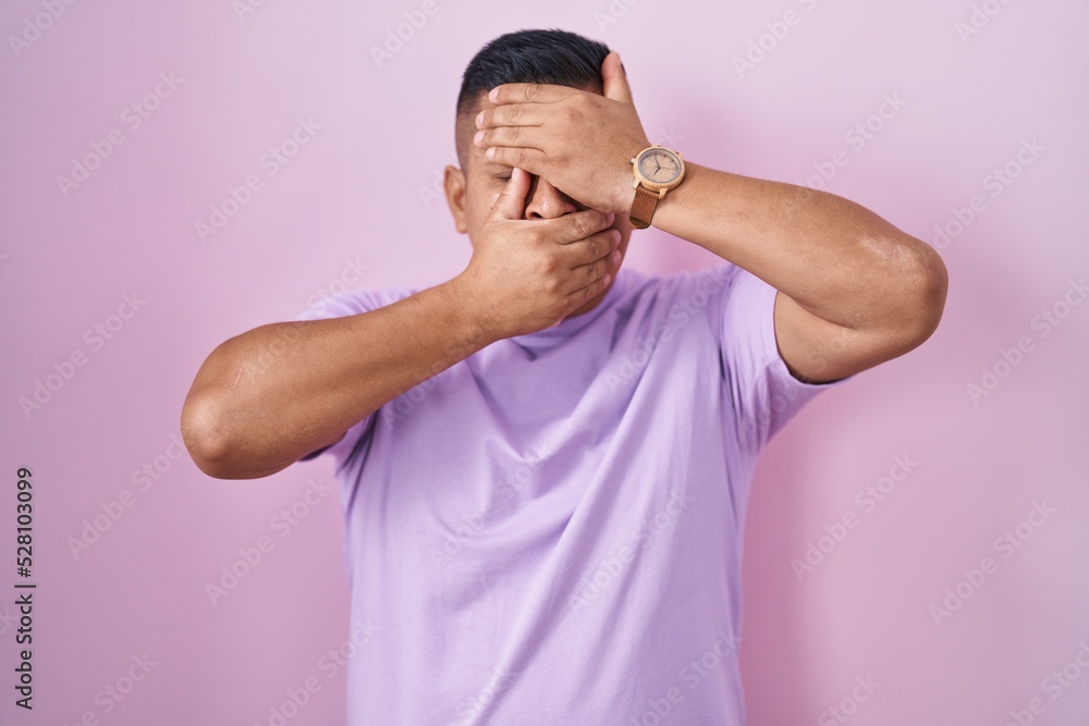 Young hispanic man standing over pink background covering eyes and mouth with hands, surprised and shocked. hiding emotion Young hispanic man standing over pink background covering eyes and mouth with hands, surprised and shocked. hiding emotion