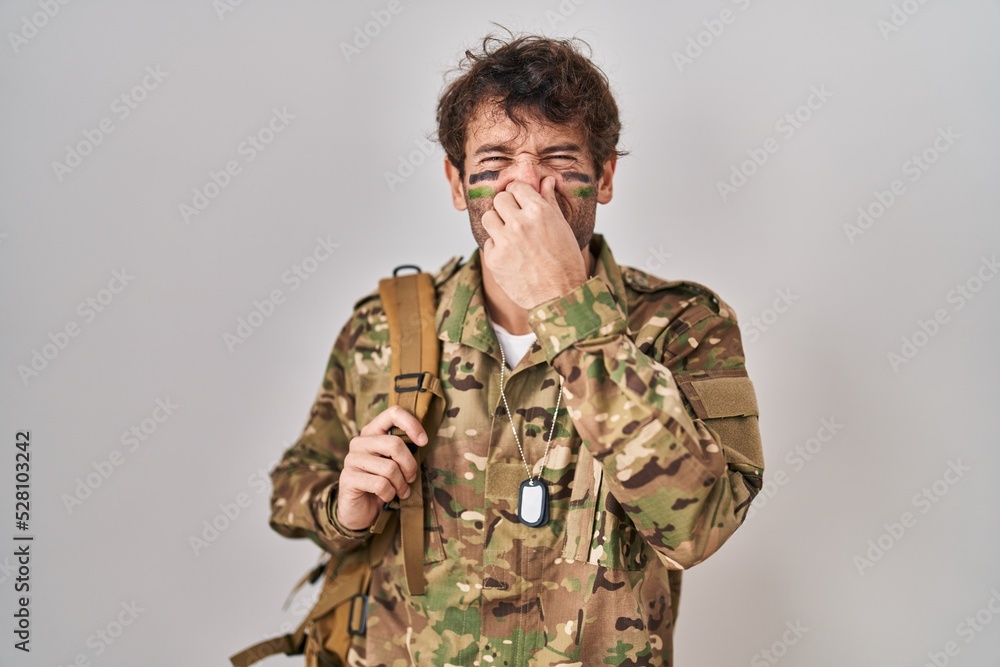 Hispanic young man wearing camouflage army uniform smelling something stinky and disgusting, intolerable smell, holding breath with fingers on nose. bad smell