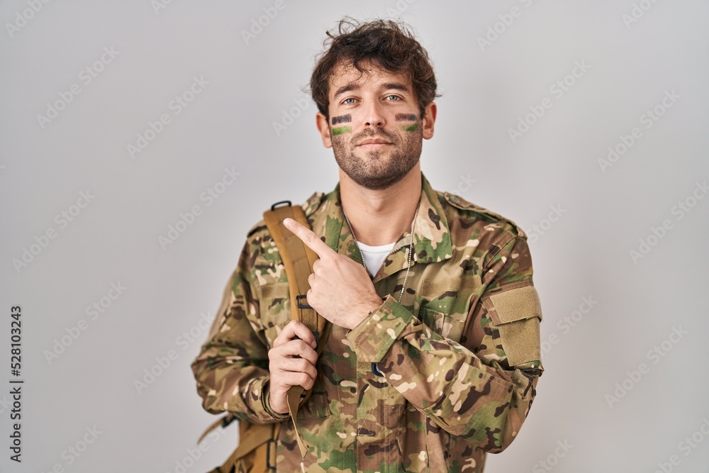 Hispanic young man wearing camouflage army uniform pointing with hand finger to the side showing advertisement, serious and calm face