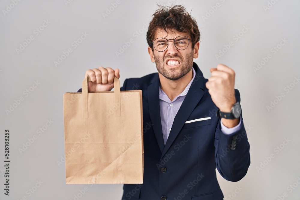 Hispanic business man holding delivery bag annoyed and frustrated shouting with anger, yelling crazy with anger and hand raised Hispanic business man holding delivery bag annoyed and frustrated shouting with anger, yelling crazy with anger and hand raised