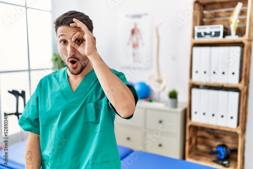 Young physiotherapist man working at pain recovery clinic doing ok gesture shocked with surprised face  eye looking through fingers. unbelieving expression.