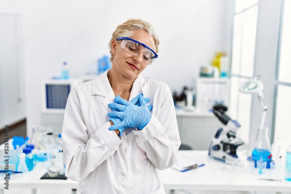 Middle age blonde woman working at scientist laboratory smiling with hands on chest with closed eyes and grateful gesture on face. health concept. Middle age blonde woman working at scientist laboratory smiling with hands on chest with closed eyes and grateful gesture on face. health concept.