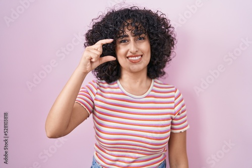 Young middle east woman standing over pink background smiling and confident gesturing with hand doing small size sign with fingers looking and the camera. measure concept.