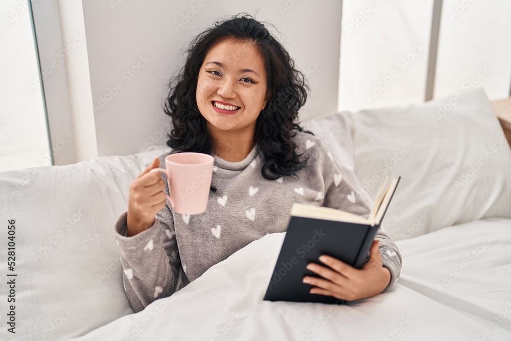 Young chinese woman dinking coffee and reading book sitting on bed at bedroom Young chinese woman dinking coffee and reading book sitting on bed at bedroom