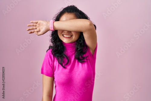 Young asian woman standing over pink background covering eyes with arm smiling cheerful and funny. blind concept.