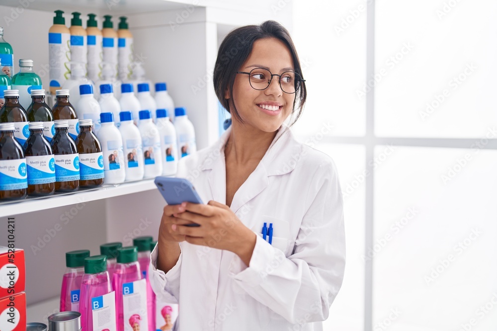 Young hispanic woman pharmacist using smartphone working at pharmacy