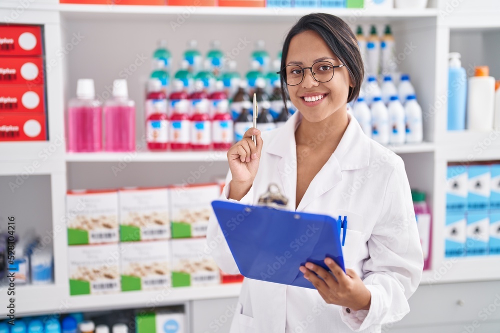 Young hispanic woman pharmacist writing on document looking shelving at pharmacy