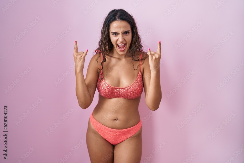 Young hispanic woman wearing lingerie over pink background shouting with crazy expression doing rock symbol with hands up. music star. heavy music concept.