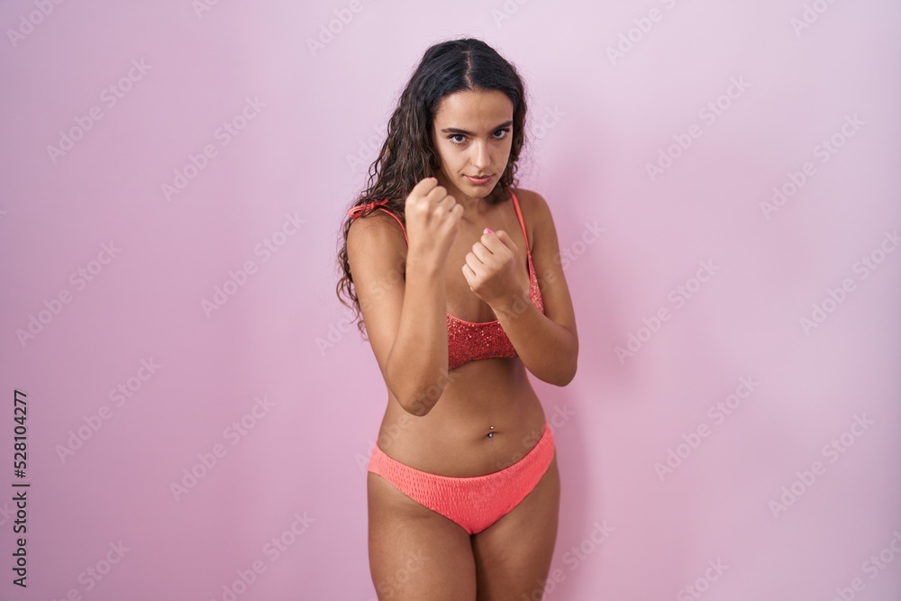 Fototapeta premium Young hispanic woman wearing lingerie over pink background ready to fight with fist defense gesture, angry and upset face, afraid of problem