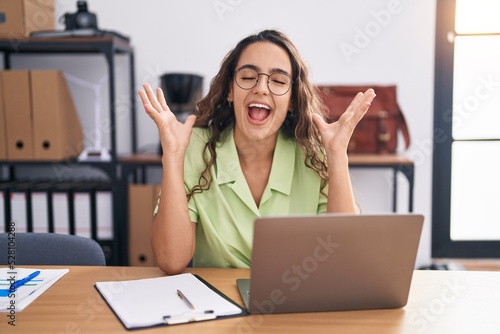 Young hispanic woman working at the office wearing glasses celebrating mad and crazy for success with arms raised and closed eyes screaming excited. winner concept