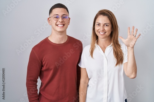 Mother and son standing together over isolated background showing and pointing up with fingers number five while smiling confident and happy.