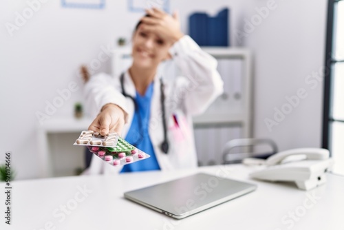 Young hispanic doctor woman wearing doctor uniform holding prescription pills at clinic stressed and frustrated with hand on head  surprised and angry face