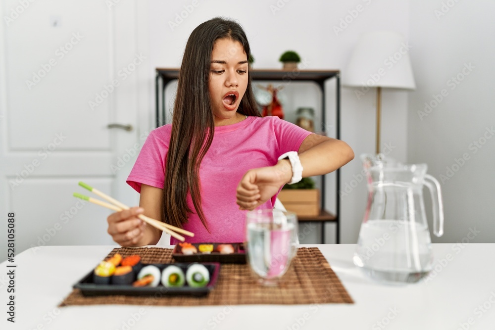 Young brunette woman eating sushi using chopsticks looking at the watch time worried, afraid of getting late Young brunette woman eating sushi using chopsticks looking at the watch time worried, afraid of getting late