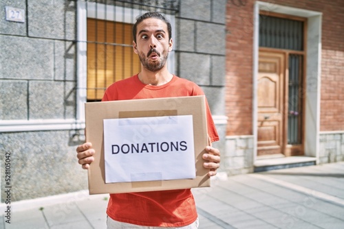 Young hispanic man holding donations box for charity outdoors making fish face with mouth and squinting eyes, crazy and comical.