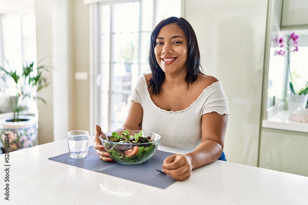 Hispanic brunette woman eating green salad at the kitchen