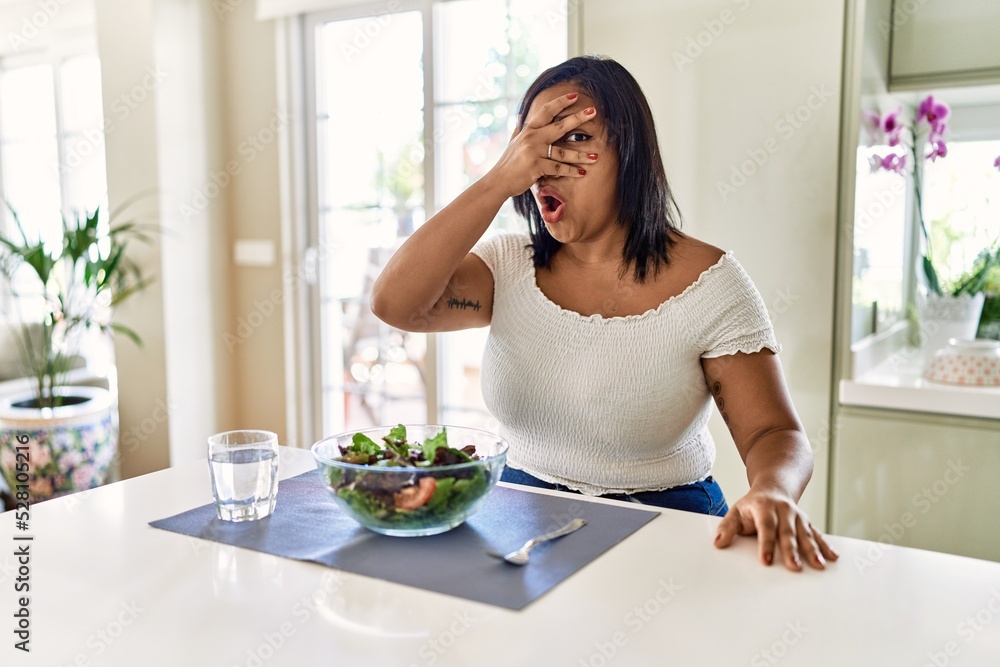 Young hispanic woman eating healthy salad at home peeking in shock covering face and eyes with hand, looking through fingers with embarrassed expression.